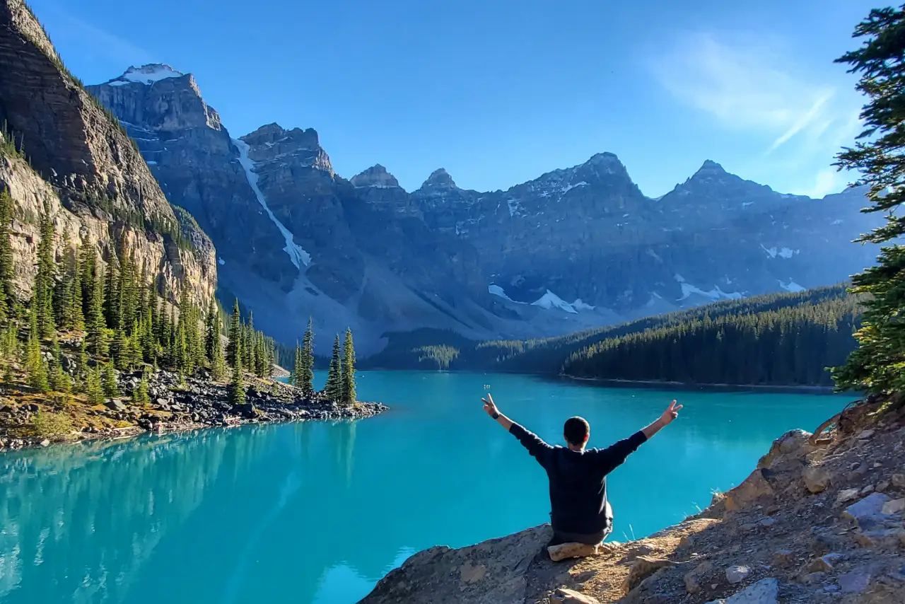 Person with raised arms sitting by a turquoise lake surrounded by mountain peaks.