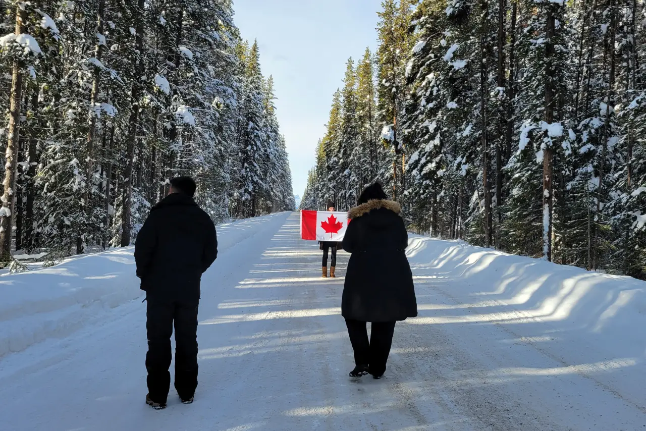 Two individuals walking on a snowy forest road, one holding a Canadian flag.