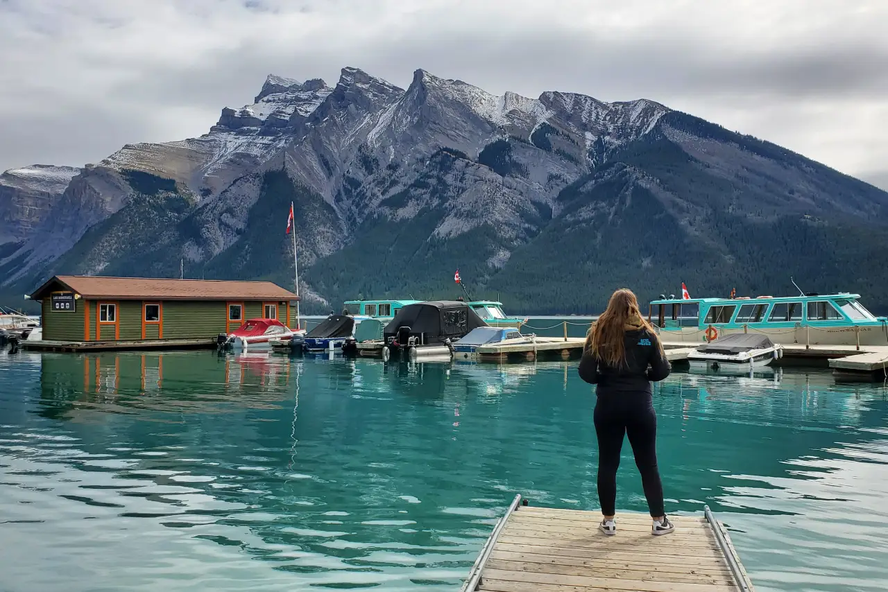 Experience Summer in Banff - summer-tours 1 Person on a dock overlooking a turquoise lake and mountain range with boats and a cabin.