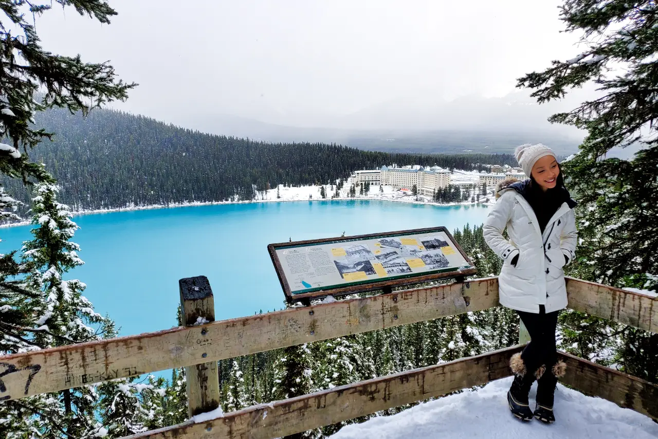 Experience Summer in Banff - summer-tours 2 A person in winter clothing stands by an information sign overlooking a turquoise lake and a large hotel.