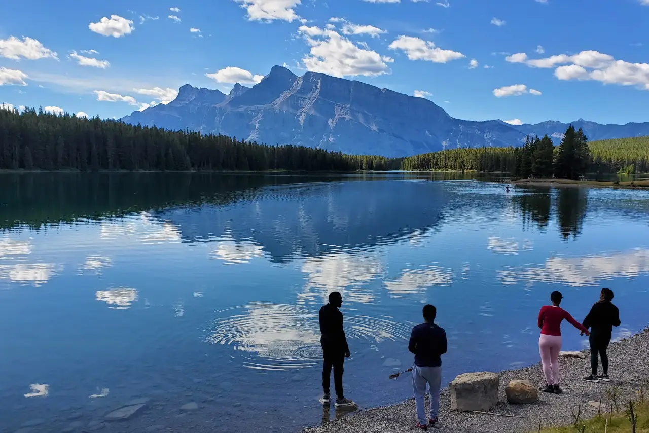 Ultimate Experience - summer-tours 1 People by a tranquil lake with a mountain backdrop and reflections of clouds on the water.