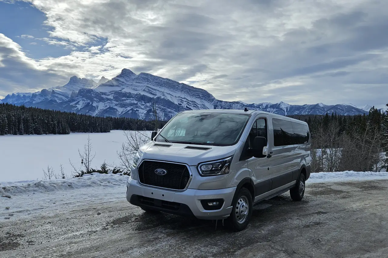 Discover Banff on a Premium Day Trip 1 A silver Ford van parked on a snowy road with a stunning backdrop of mountains and a frozen lake under a cloudy sky.