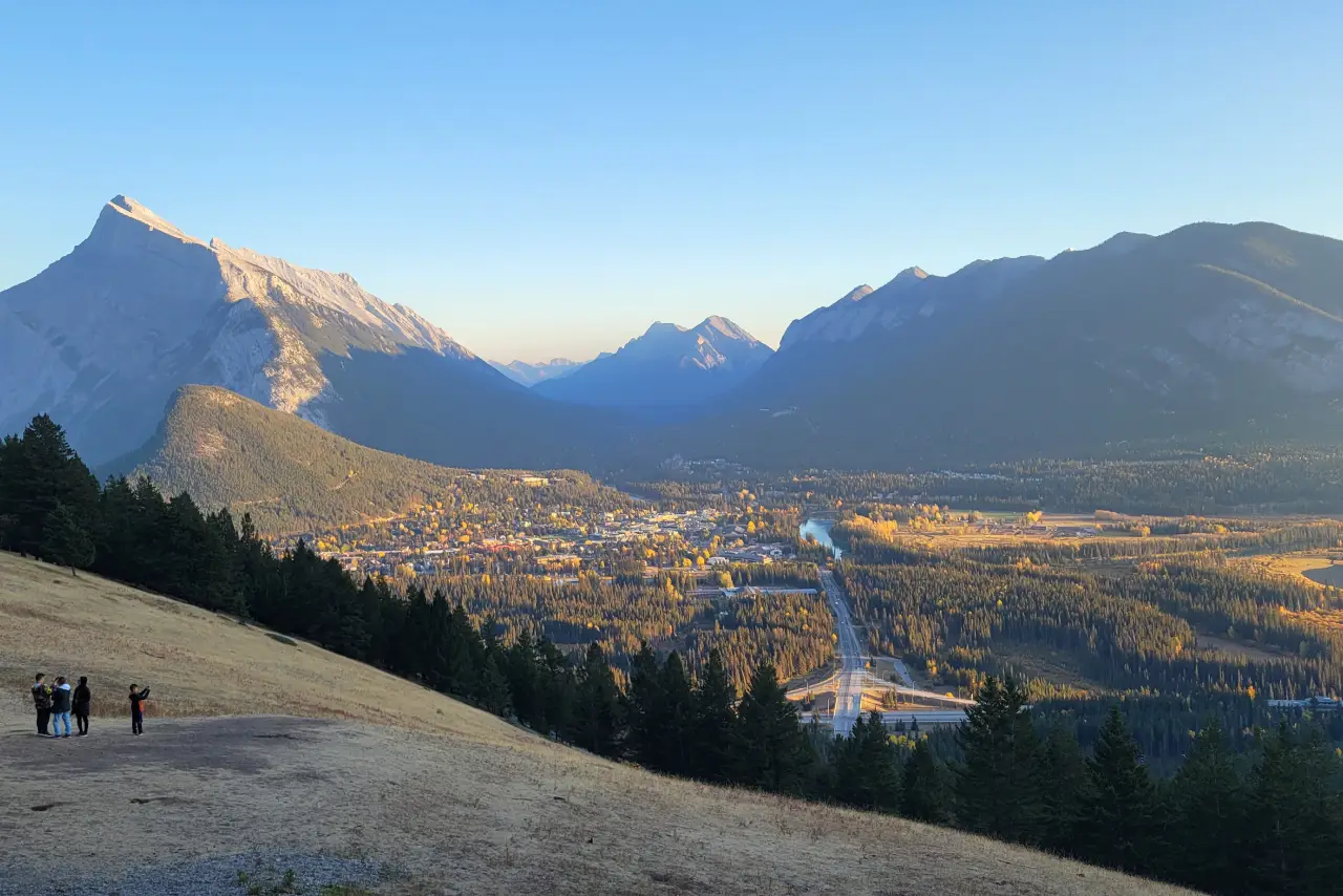 Explore Banff’s Iconic Beauty on a Full-Day Adventure A breathtaking view of mountains surrounding a valley town, with a winding river and autumn foliage under a clear blue sky.