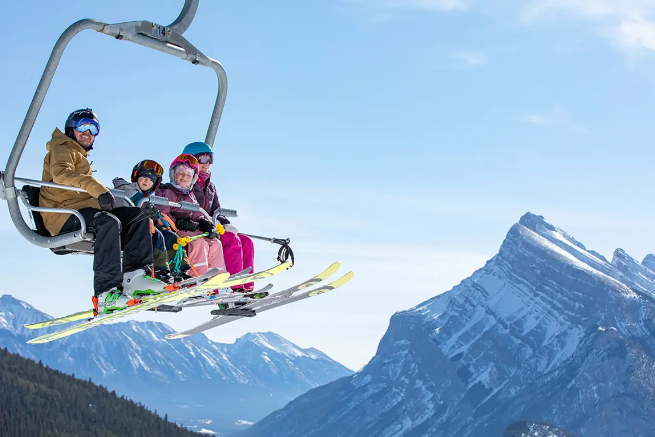 Join Us on a Wild Adventure 1 A group of four people on a ski lift with colorful gear, surrounded by snowy mountains and clear blue skies.