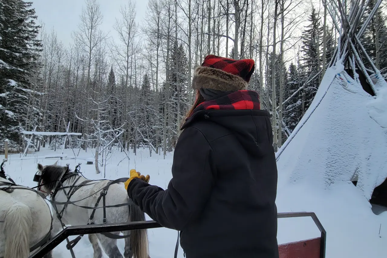 Join Us on a Wild Adventure A person in winter gear stands beside a sled, looking towards a snowy landscape with horses and a teepee in the background.